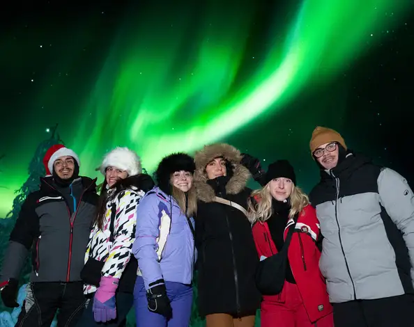 A group of seven guests smiling under a bright green aurora on an Experience Tours Northern Lights hunt near Rovaniemi